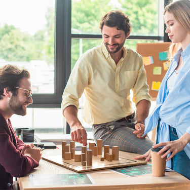 In einem Startup wird von zwei Männern und einer Frau eine Aufstellung gemacht. Die Frau steht, ein Mann sitz auf einem Stuhl und ein Mann sitzt lässig auf dem Tisch. Sie planen strategisch mit dem Pro Systembrett